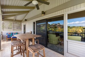 A covered patio with a wooden table and chairs, a bench with colorful cushions, and a view of a residential area through a glass door.