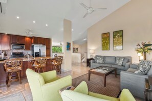 Eine moderne Küche mit Holzschränken, ein Essbereich mit Korbstühlen und ein Wohnzimmer mit grauem Sofa und grünen Sesseln.