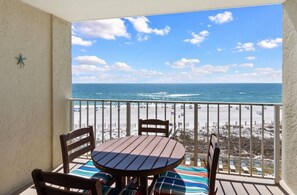 Un balcon avec une table et des chaises en bois, donnant sur une plage, avec un ciel dégagé et une vue sur l’océan.