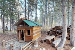 A rustic wooden cabin with a green roof, surrounded by a forest. There is a stack of firewood and a picnic table nearby.