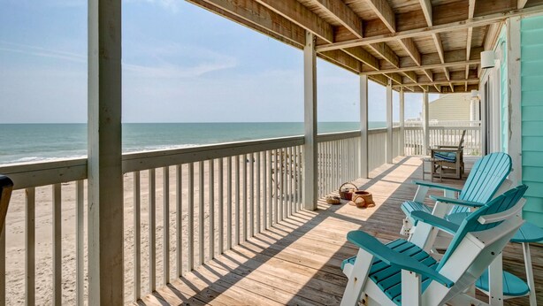 A wooden deck with a railing overlooking the ocean, featuring several blue and white Adirondack chairs and a small table with baskets.