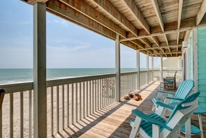 Een houten terras met een leuning, met uitzicht op de oceaan, met blauw-witte Adirondack-stoelen en een kleine tafel met manden.