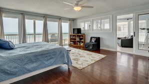 A bedroom with a bed, a ceiling fan, a bookshelf, a small table, and a view of the ocean.