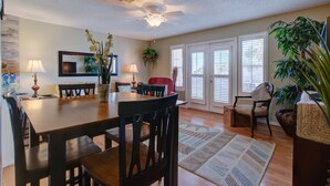 A dining area with a wooden table, chairs, and a ceiling fan. There are potted plants and a rug on the floor.