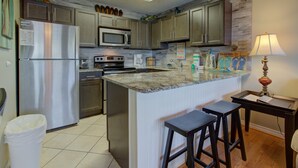 A kitchen with a stainless steel refrigerator, a microwave, and a sink. There are wooden cabinets, a marble countertop, and two stools.