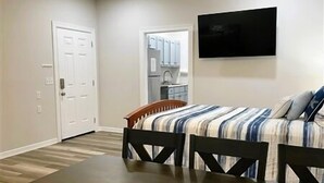 Un dormitorio con cama, televisor montado en la pared y cocina visible a través de una puerta abierta.
