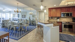 Una cocina moderna con electrodomésticos de acero inoxidable, una zona de comedor con una mesa y sillas de vidrio, y un tapete azul estampado.