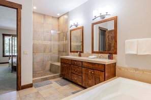A modern bathroom with a walk-in shower, a large bathtub, wooden cabinetry, and a mirror with lights.