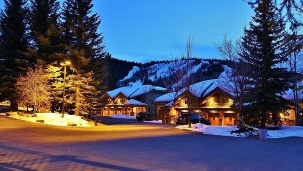 A row of log cabin-style buildings with snow-covered roofs, surrounded by tall evergreen trees and a mountain backdrop. - Indulge in Luxury - Perfect for Groups with Private Hot Tub & Posturepedic Beds! (Park City)