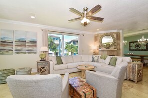 A living room with a ceiling fan, a large window with curtains, a sofa, a coffee table, and a decorative mirror. - Three-story home w/private pool, hot tub, game room & more in Destin! (Destin)