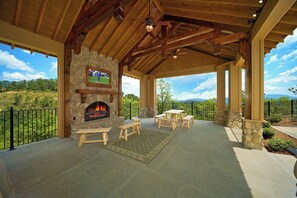 A covered patio with a stone fireplace, wooden benches, and tables, overlooking a scenic landscape.