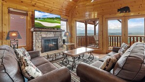 A cozy cabin interior with a stone fireplace, leather sofas, a flat-screen TV, and a scenic view through large windows. - Stunning Mountain Views | Game Room | Pet Friendly | Hot Tub | WiFi (SEVIERVILLE)