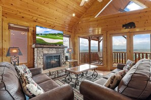A cozy cabin interior with a stone fireplace, leather seating, a television, and a scenic view through the windows. - Stunning Mountain Views | Game Room | Pet Friendly | Hot Tub | WiFi (SEVIERVILLE)