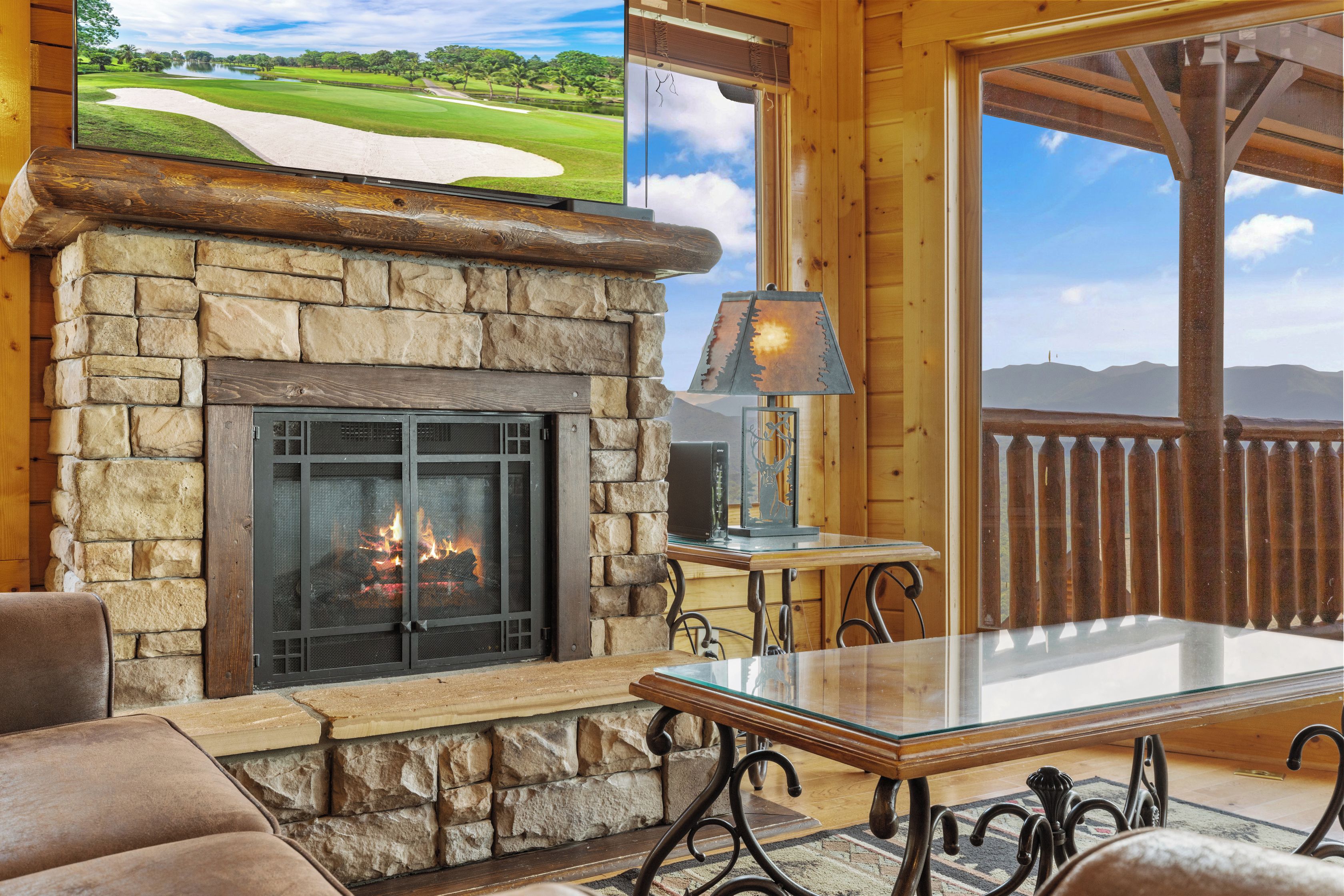 Une cheminée en pierre avec un feu de bois, une table en verre et une vue sur un terrain de golf, visible à travers une fenêtre.