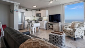 A modern kitchen with white cabinets, a microwave, and a TV. There is a dining area with a white table and chairs, and a sofa in the living room. A large window offers a view of the ocean. - Spectacular Deluxe Estero Beach & Tennis Beachfront Condo w/ Direct Gulf View (Fort Myers Beach)