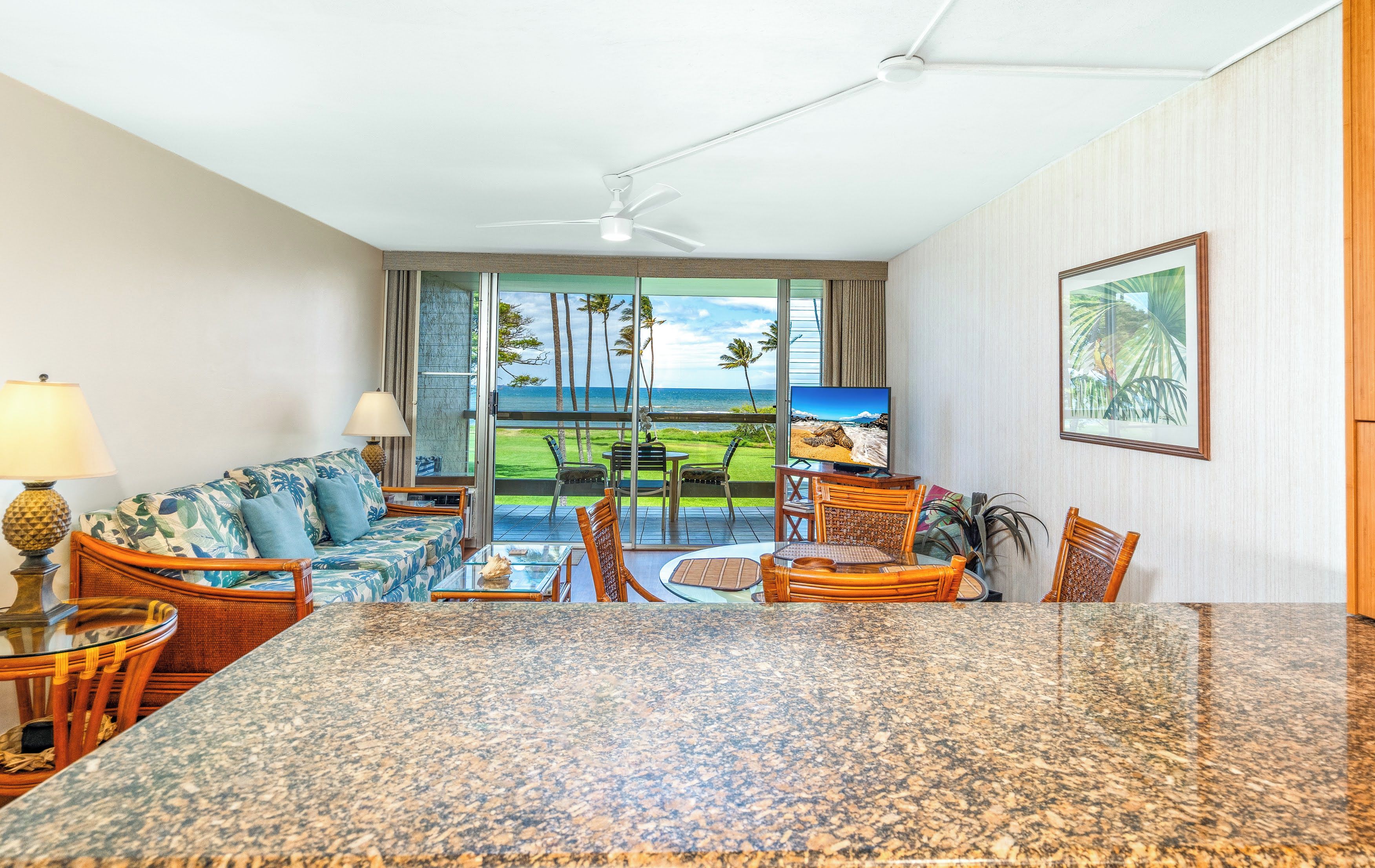 A hotel room with a large granite table, a sofa with tropical cushions, a television, and a view of the ocean through large sliding doors.