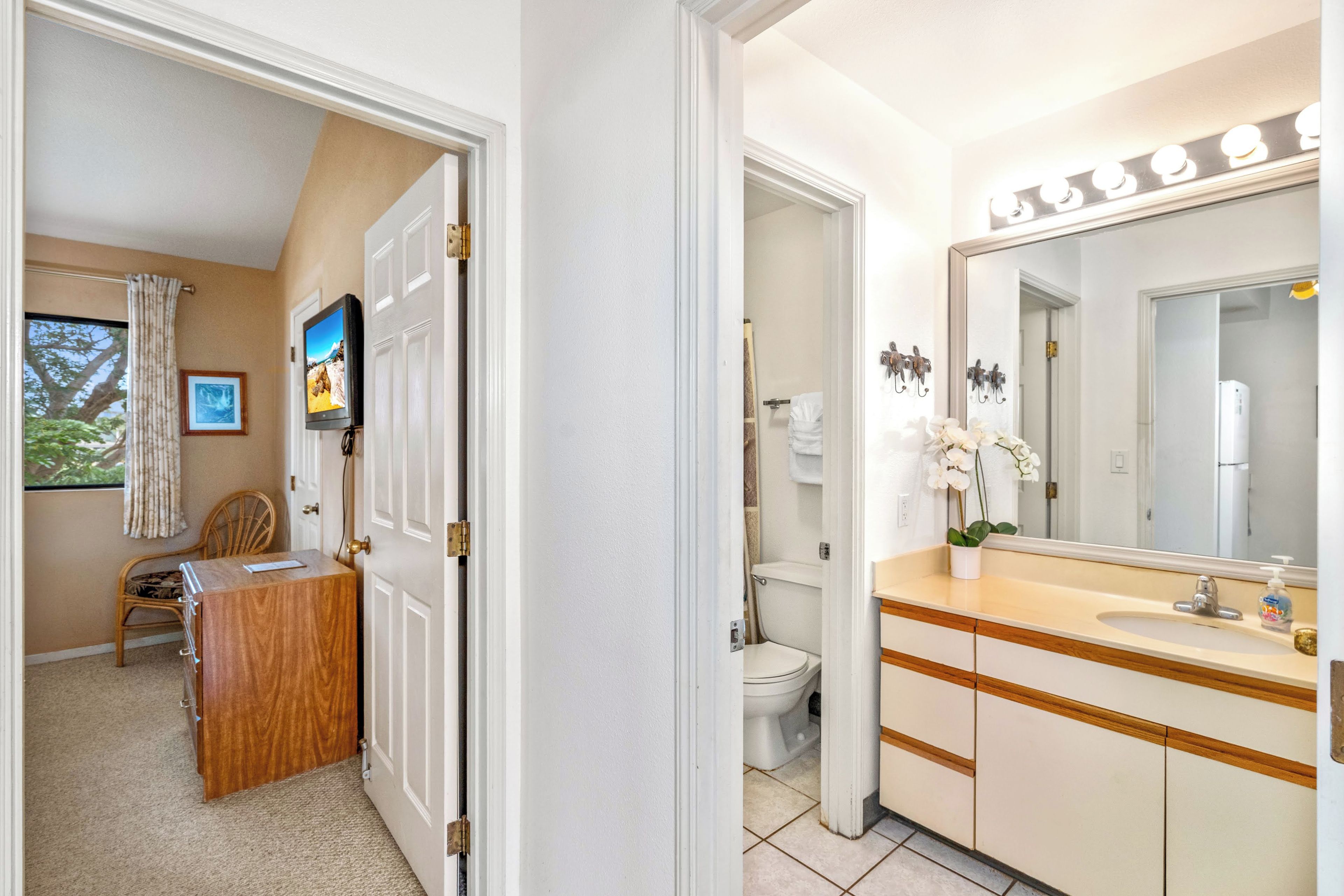 Un baño con un espejo grande, lavabo e inodoro. En la habitación contigua hay un aparador de madera y una silla.