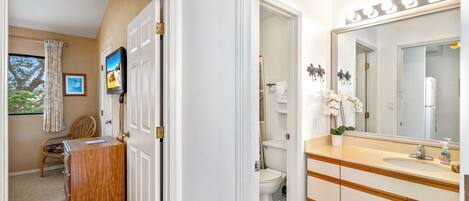 Un baño con un espejo grande, lavabo e inodoro. En la habitación contigua hay un aparador de madera y una silla.