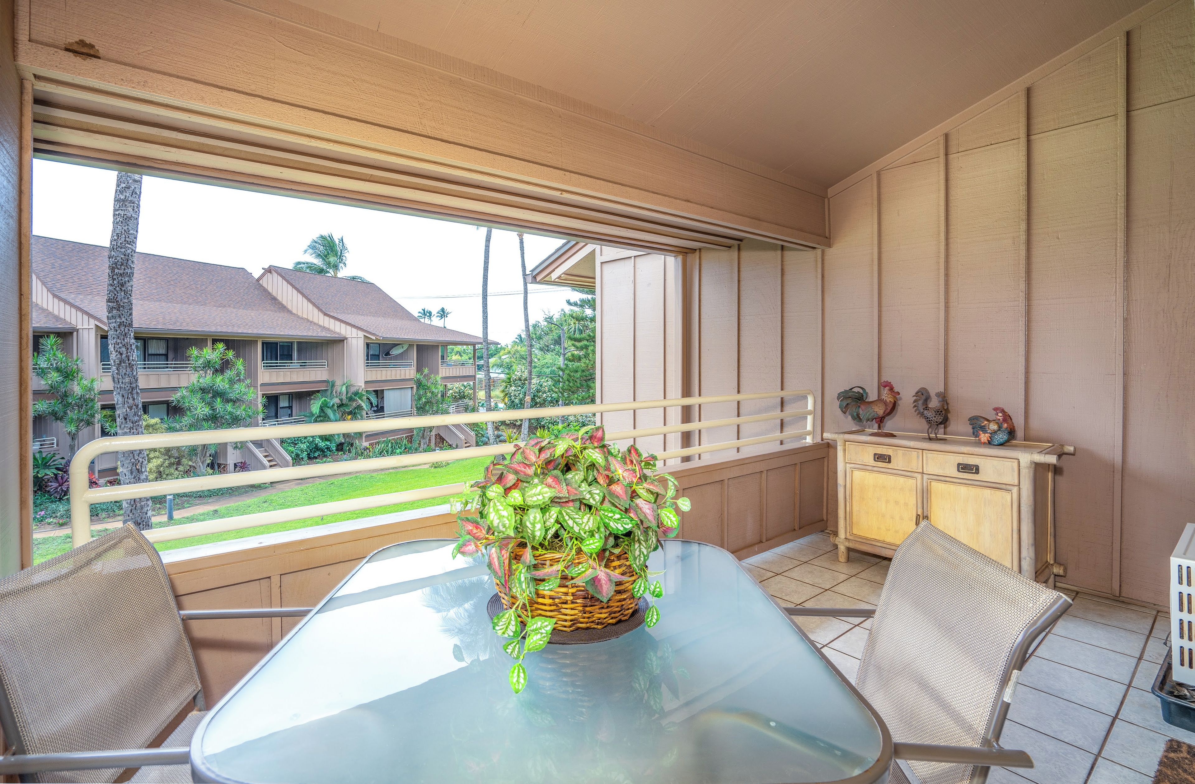 A balcony with a glass table, wicker chairs, a wicker basket with plants, and a wooden cabinet with decorative items.