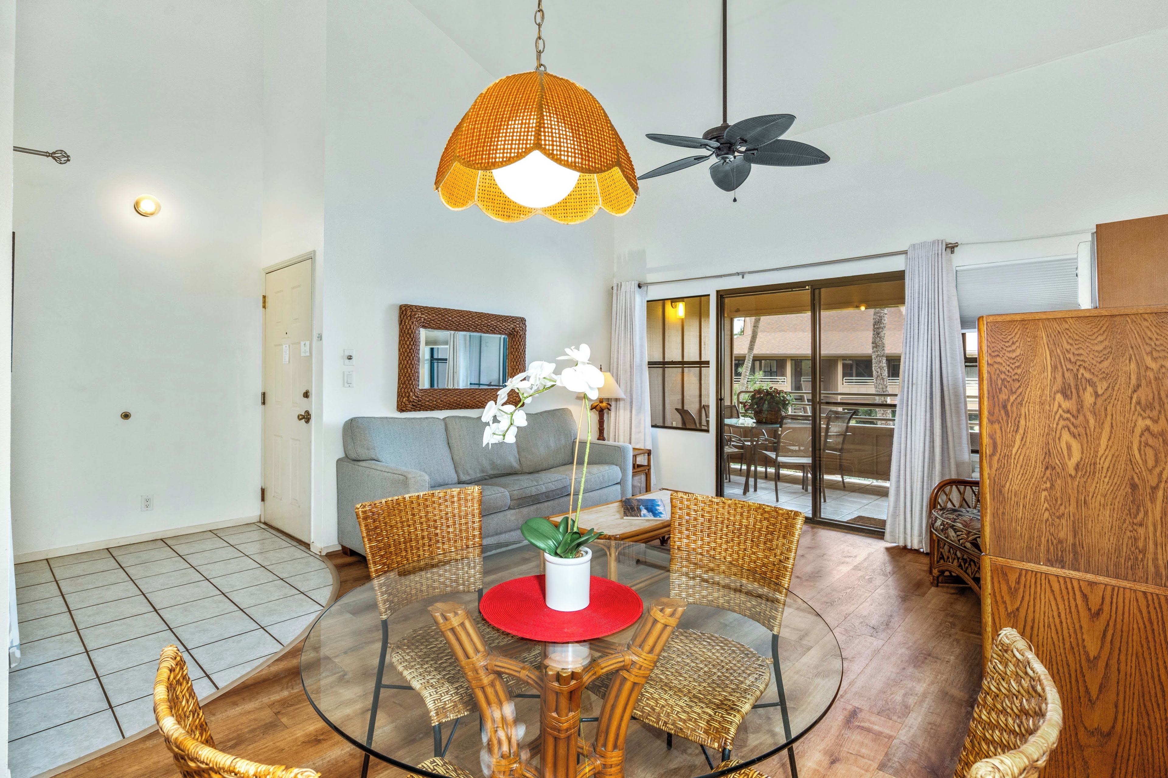 A spacious living area with a glass dining table surrounded by wicker chairs, a grey sofa, and a ceiling fan.