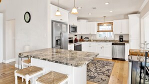 A modern kitchen with white cabinets, stainless steel appliances, a granite countertop, and a dining area with white stools. - Gulf Views | Beach Access | Pet Friendly Coastal Bungalow (Gulf Shores)