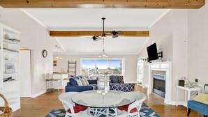 A spacious living room with a glass-top dining table surrounded by wicker chairs, a fireplace, and a large window offering a view of the outdoors. - Gulf Views | Beach Access | Pet Friendly Coastal Bungalow (Gulf Shores)