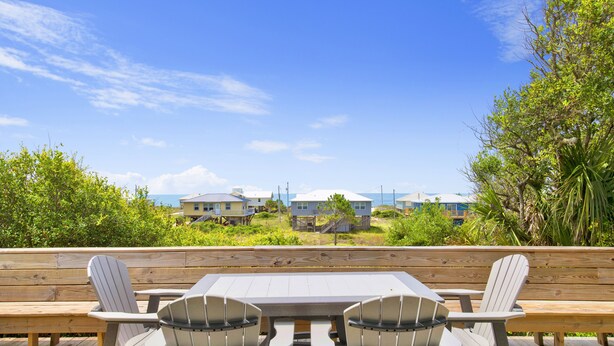 A wooden deck with a table and chairs, overlooking a beachfront property with houses and greenery. - Gulf Views | Beach Access | Pet Friendly Coastal Bungalow (Gulf Shores)