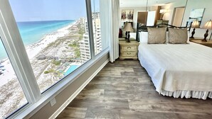 Ein Hotelzimmer mit großem Fenster mit Blick auf Strand und Meer, einem Bett mit weißer Bettwäsche, einem Nachttisch mit Lampe und einem Spiegel.