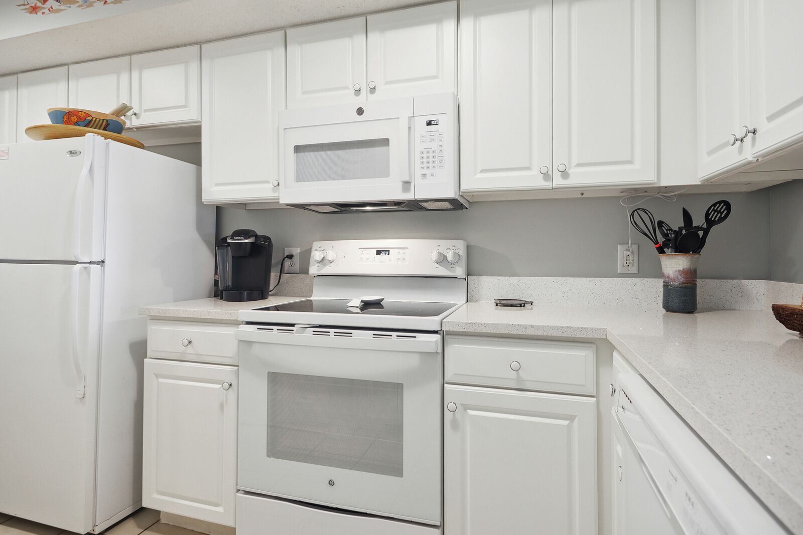 Una cocina con armarios blancos, un refrigerador, un microondas, una cafetera y una estufa.