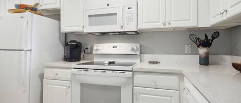 Una cocina con armarios blancos, un refrigerador, un microondas, una cafetera y una estufa.