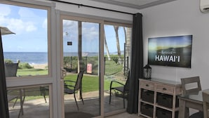 A room with a sliding glass door leading to a balcony with a view of the ocean and palm trees. There is a flat-screen TV mounted on the wall and a small wooden desk with drawers.