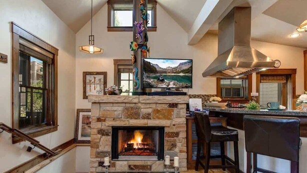 A modern kitchen with a stone fireplace, a dining area with bar stools, and a large window overlooking a scenic landscape.
