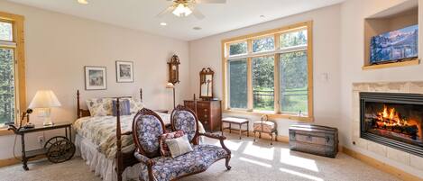 A spacious bedroom with a large bed, a classic chair, a fireplace, and a large window providing natural light.