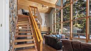 Una escalera de madera con un muro de piedra, un ventanal con vistas a árboles y una zona de estar acogedora con un tapete estampado.
