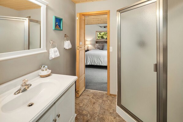 Un baño con un espejo grande, un lavamanos y una puerta de madera que da a un dormitorio con una cama y una mesita de noche.