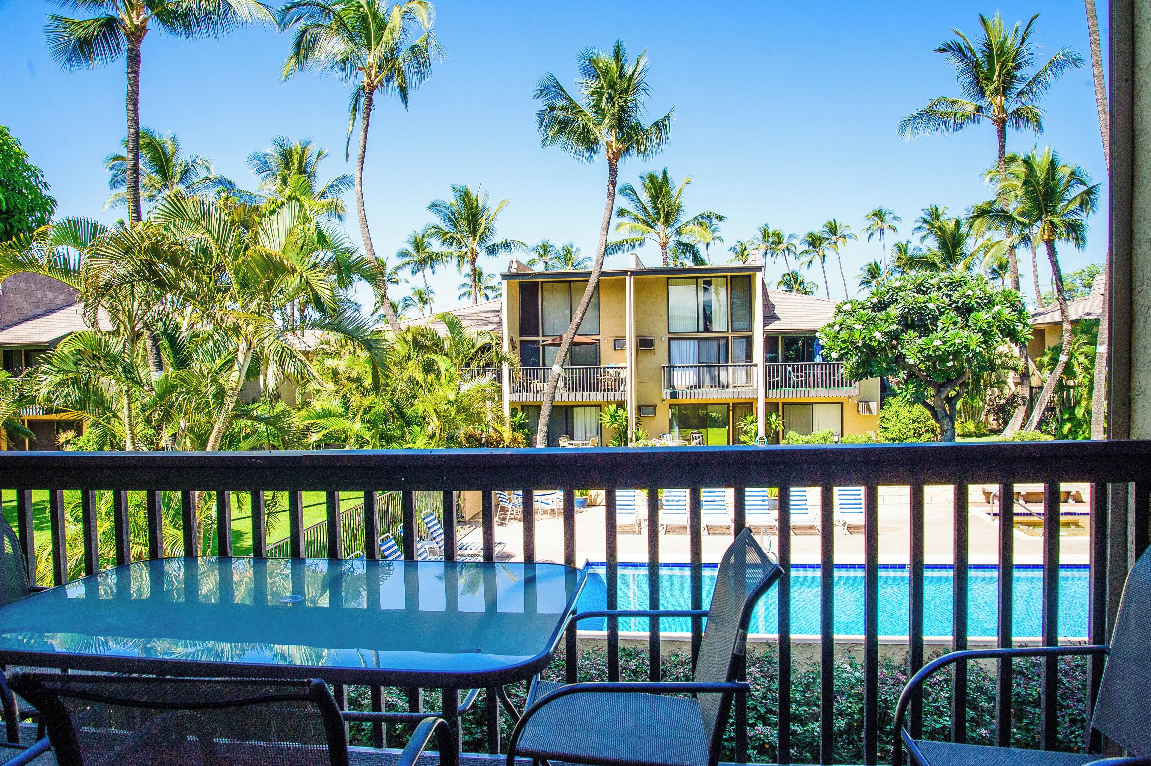 A balcony with a glass table and chairs overlooking a swimming pool, surrounded by palm trees and a building with a balcony.