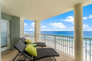 A balcony with two wicker chairs and a table, overlooking a beach with umbrellas and people.