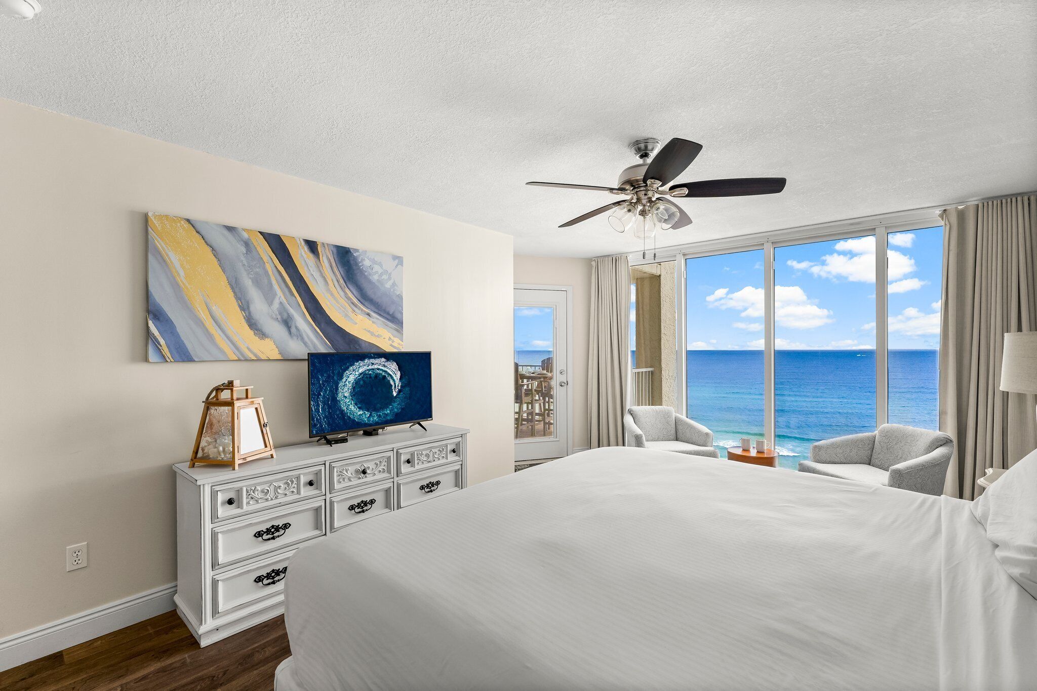 Habitación de hotel con una cama grande, ventilador de techo, televisión, cómoda y vista al océano.