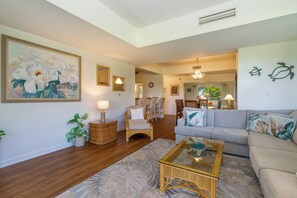 A living room with a sofa, a coffee table, a chair, a floor lamp, and a painting on the wall. - Vista Waikoloa Condo w/ Mountain Views - Walk to Beach! (Waikoloa)