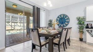A dining area with a large clock, chairs, a table set for four, and a view of the outside through a sliding door. - 5 bed with spa and games room (Davenport)