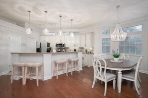 Cozinha com armários brancos, uma ilha central com banquetas, uma mesa de jantar com cadeiras e iluminação pendente.