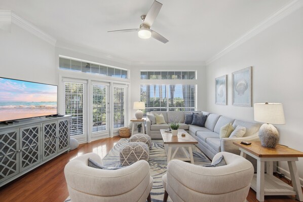 A spacious living room with a large TV, comfortable seating, and a view of the outdoors through large windows. - 3BR Beach Villa Near Pool! New 6-Seater GOLF CART, Screened Porch, Beach Tram (Miramar Beach)