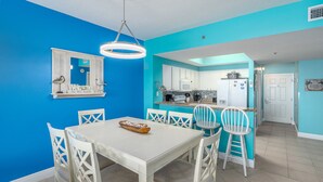 A dining area with a white table and chairs, a kitchen with white cabinets and a blue countertop, and a bright blue wall. - After Dune Delight ~ 2BR/2BA ~ 15th Floor ~ (Miramar Beach)