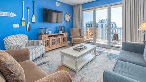 A living room with a blue wall, a flat-screen TV, a clock, and a view of the city through large windows. - After Dune Delight ~ 2BR/2BA ~ 15th Floor ~ (Miramar Beach)