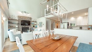 Amplia sala de estar con mesa de comedor de madera, sillas blancas, chimenea y cocina al fondo.