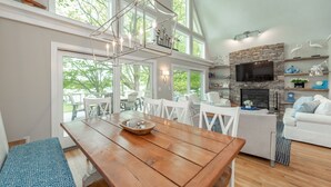 Un amplio salón con una mesa de comedor de madera, muebles blancos, una chimenea de piedra y ventanas grandes que ofrecen vistas a la vegetación.