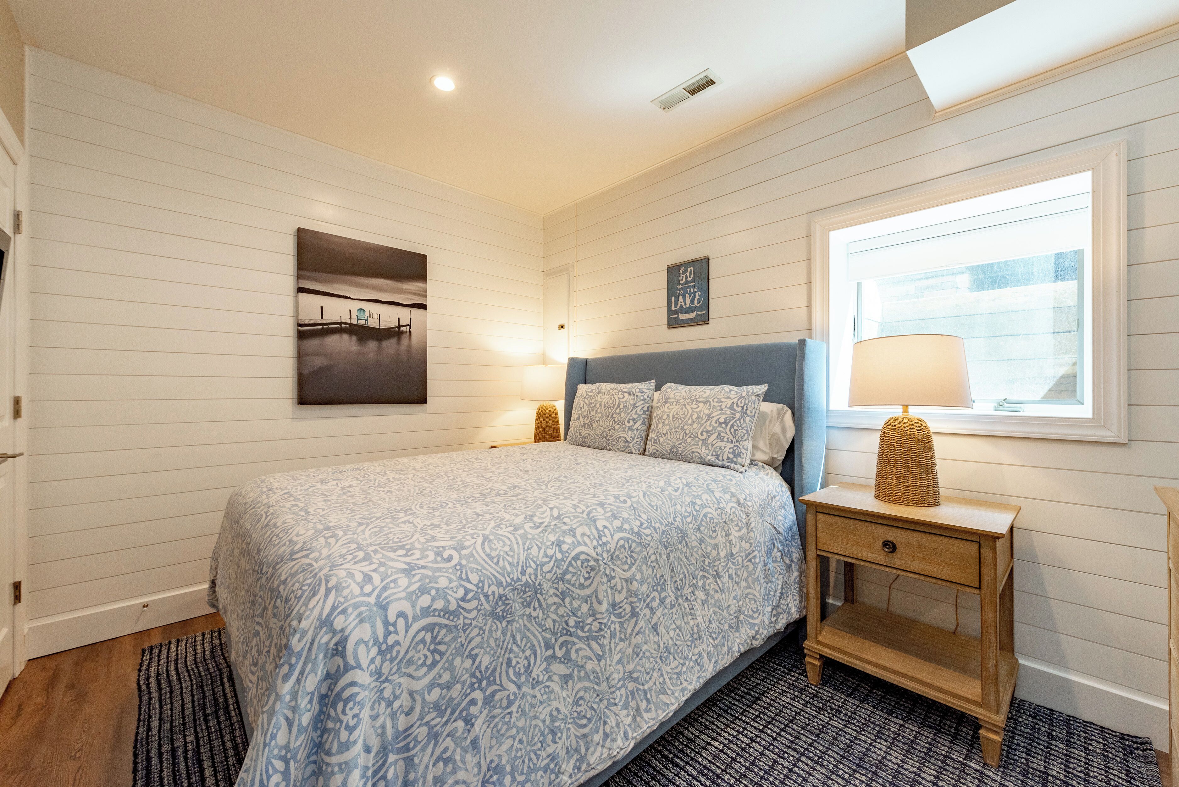 A bedroom with a bed, a nightstand, a lamp, a picture on the wall, and a window.