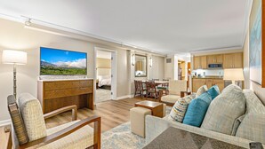 A modern living room with a flat-screen TV, a sofa with patterned cushions, a dining area with a table and chairs, and a kitchen with wooden cabinets and a microwave.