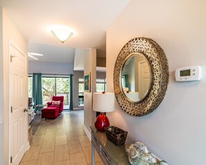 A hotel hallway with a red chair, a mirror, a thermostat, and a view of the outside.