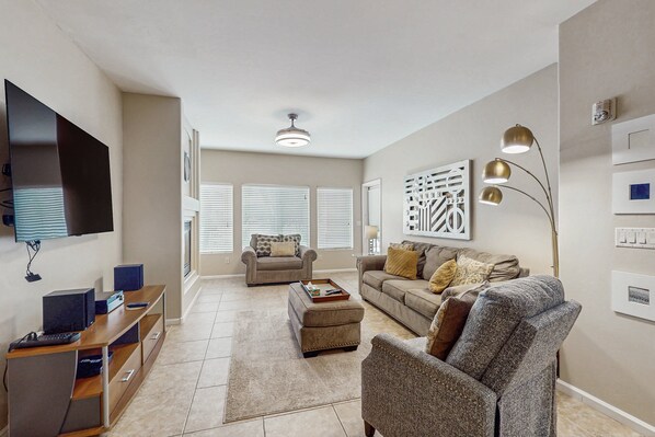A spacious living room with a flat-screen TV, a wooden entertainment unit, a large ottoman, and a gray armchair.